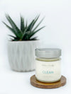 A picture of clean (ocean mist, sea grass, agave nectar and coconut milk) soy wax candle on a brown wooden coaster in front of a house plant.